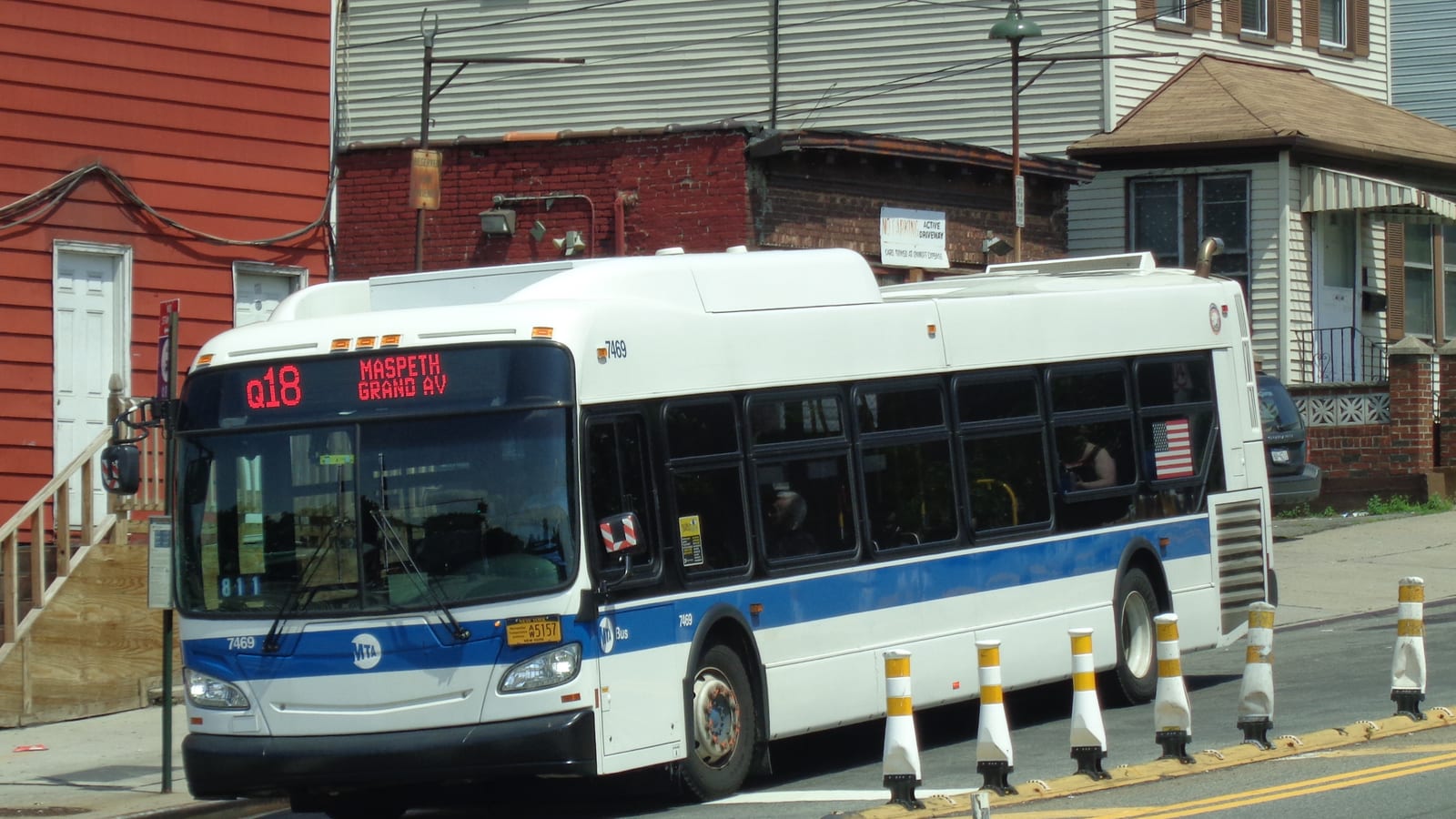 A white and blue MTA bus with the sign Q18 Maspeth Grand Av turning on a street beside a row of yellow and white traffic posts, with a red clapboard house with steps at left, a low red brick building with a sign about an active driveway, and grey sided houses behind