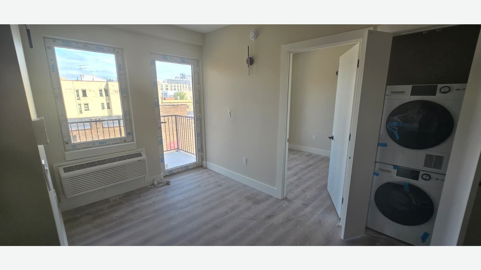 Photograph of an empty room with grey plank floors, a window and a glass balcony door with protective film, a wall air-conditioning unit, an open door to a second room, and a stacked white washer and dryer in a closet on the right