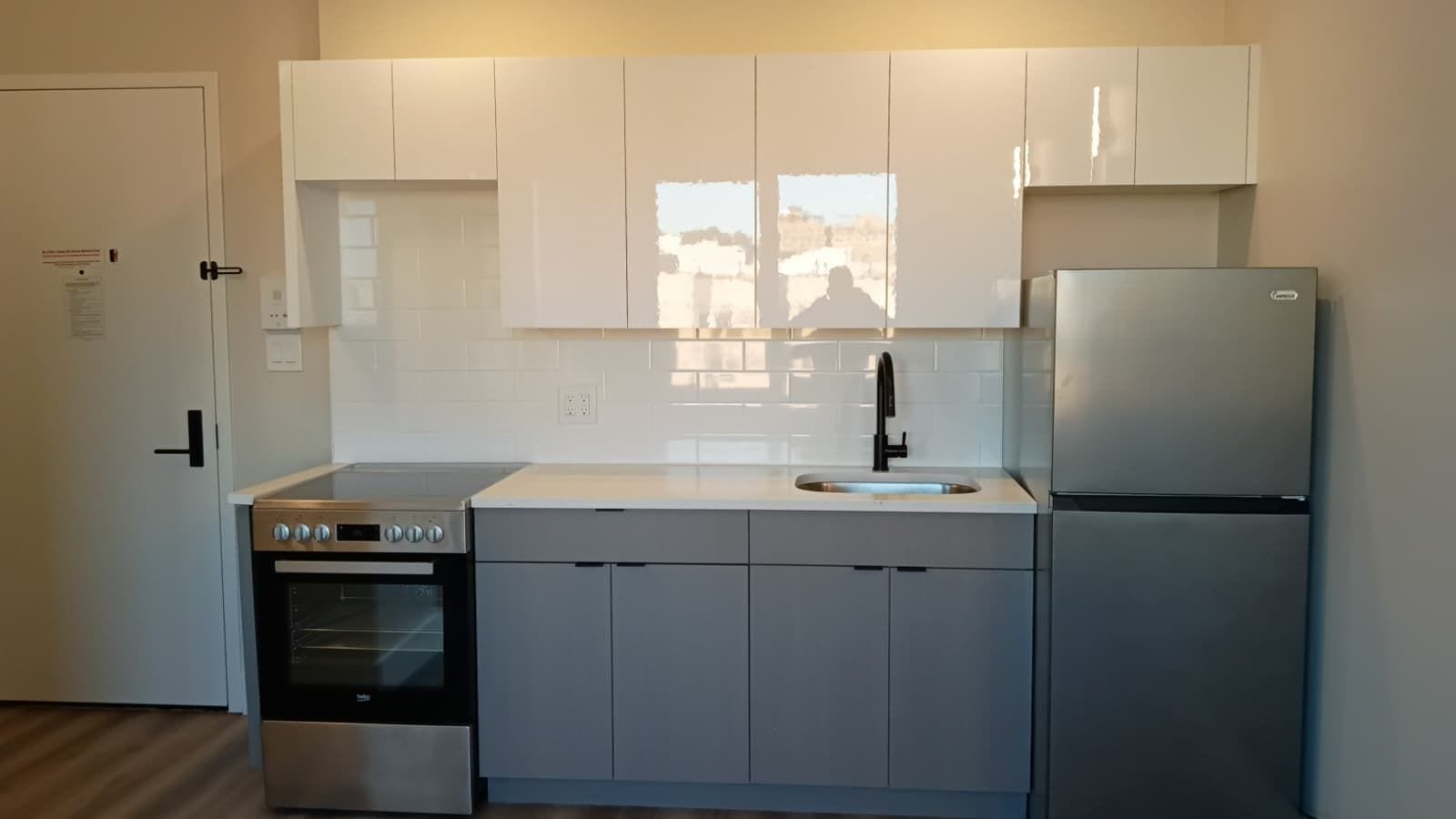 Photograph of a kitchen wall with glossy white upper cabinets, a white subway-tile backsplash, grey lower cabinets, a stainless electric range with a smooth glass top, a black faucet and a stainless refrigerator, with the apartment door on the left