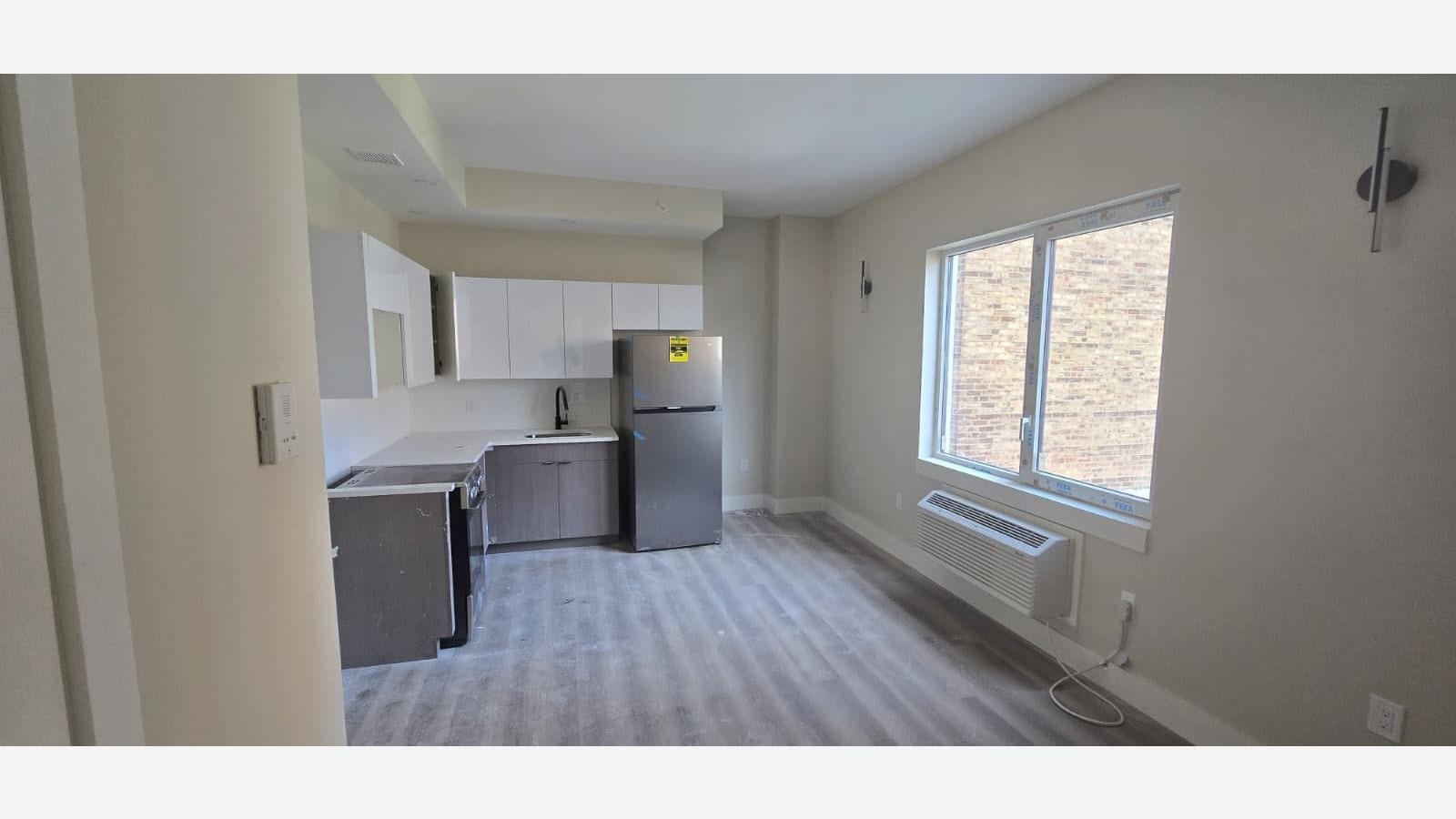 Photograph of an open room with grey plank floors, a corner kitchen with white upper and brown lower cabinets, a stainless refrigerator still taped, a window with protective film over a wall air-conditioning unit, and two wall lamps