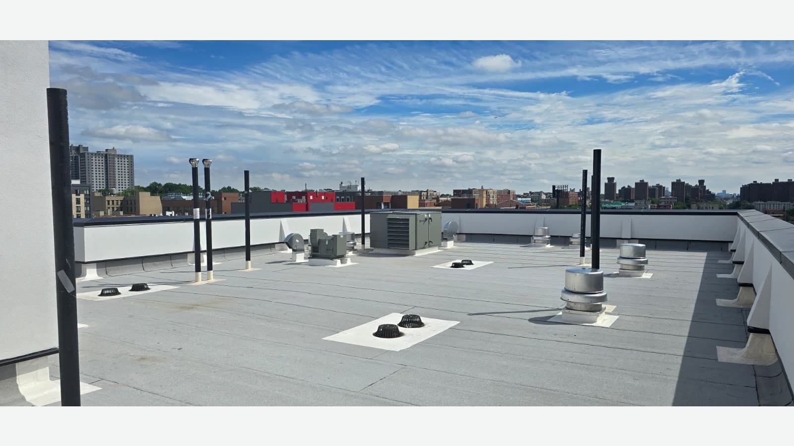 Photograph of a flat grey roof with black vent pipes, drains, metal exhaust fans and a grey mechanical unit, a white parapet wall, and Bronx apartment towers under a blue sky with thin clouds