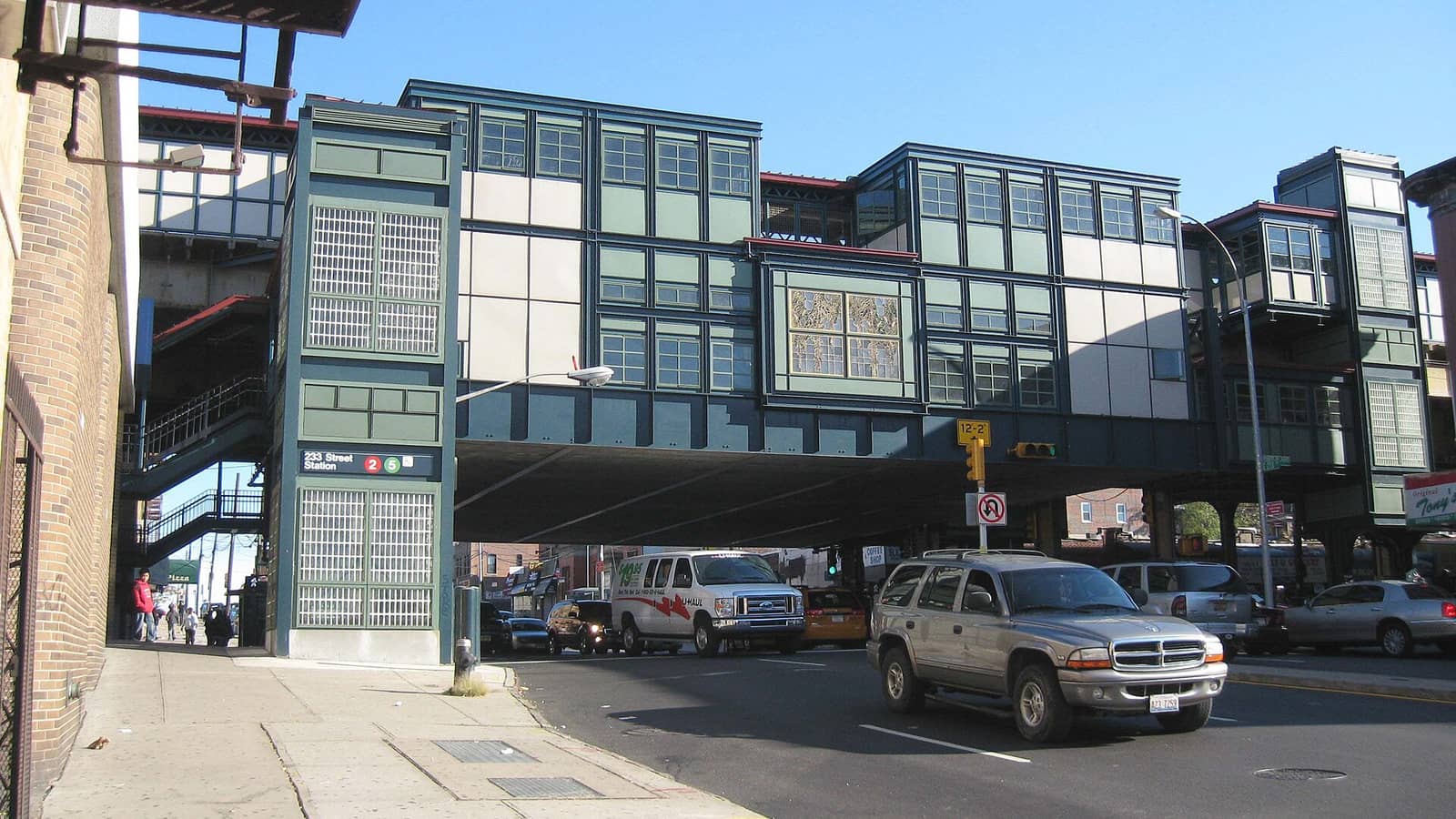 Photograph of a raised subway station built over a wide street, with green and cream paneled walls and windows, a stair tower with a sign reading 233 Street Station with red 2 and green 5 symbols, a traffic light, and a van and cars on the road below