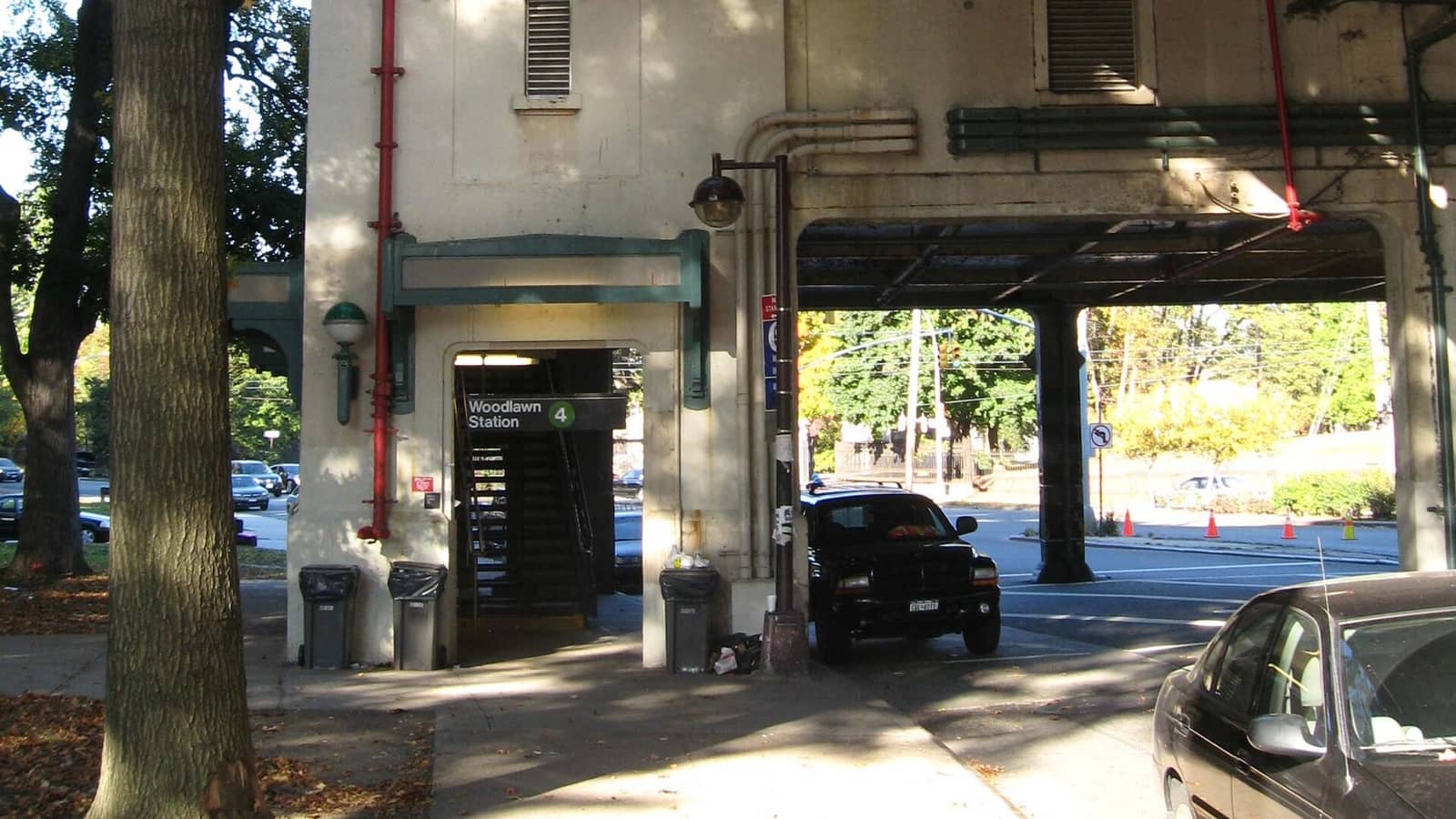 Photograph of a cream stucco stair tower under a raised subway structure, with a green-trimmed entrance and a sign reading Woodlawn Station with a green 4, red pipes on the wall, trash cans, a street lamp, parked cars and a tree trunk in the foreground