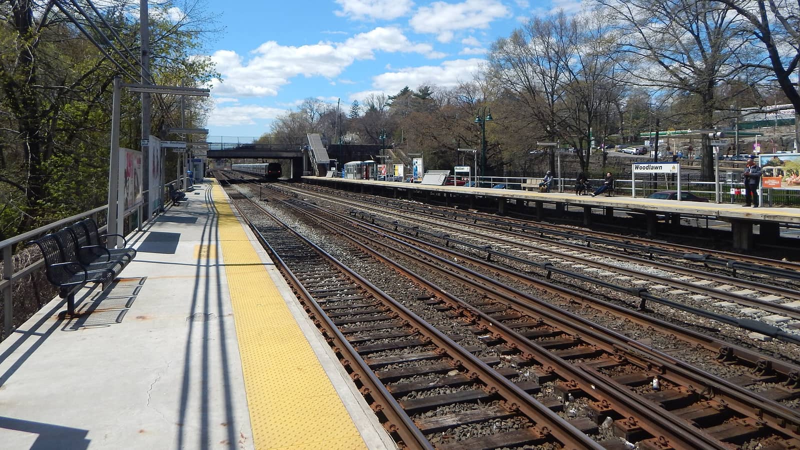 Photograph of a commuter rail station with four tracks between two long platforms, a yellow platform edge and black benches at left, a station sign reading Woodlawn at right, a footbridge and road bridge in the distance and bare and leafy trees under a blue sky