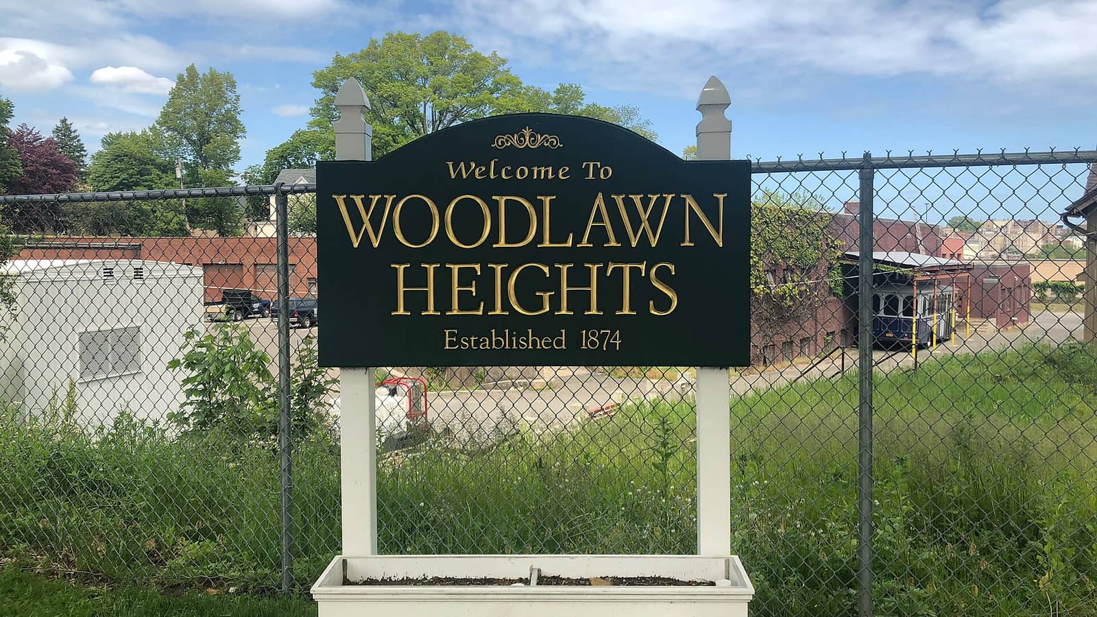 Photograph of a dark green wooden sign with gold letters reading Welcome To Woodlawn Heights, Established 1874, on white posts above a planter box, in front of a chain-link fence, tall grass, low brick buildings and trees under a partly cloudy sky