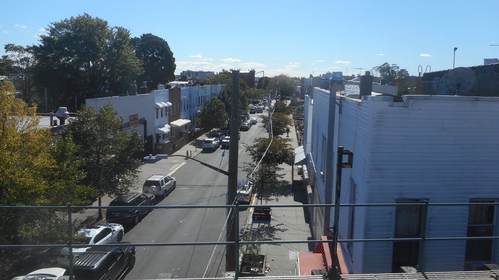 Photograph from an elevated subway platform looking down a two-way street lined with two- and three-story row houses, parked cars and street trees, with a white sided building at right and a railing in the foreground