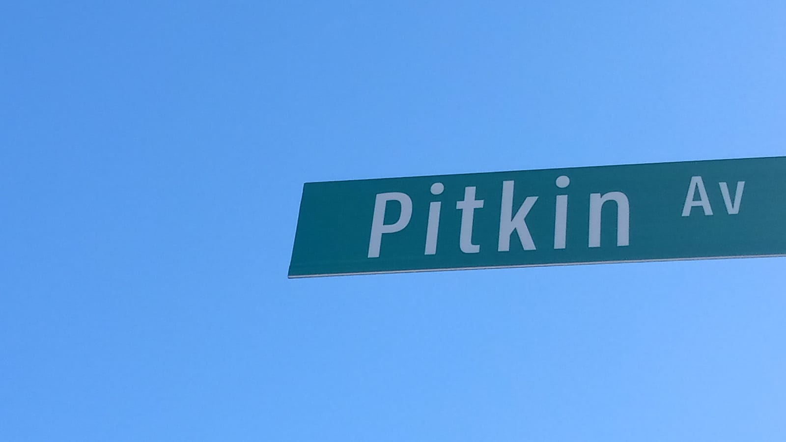 Photograph of a green street sign reading Pitkin Av against a clear blue sky