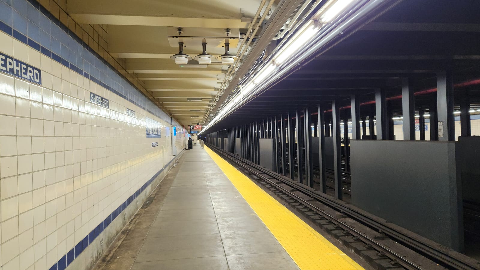 Photograph of a long, empty subway platform with tan and white wall tiles, blue Shepherd name tiles, a yellow edge strip, lamps on the ceiling and dark steel beams between the tracks