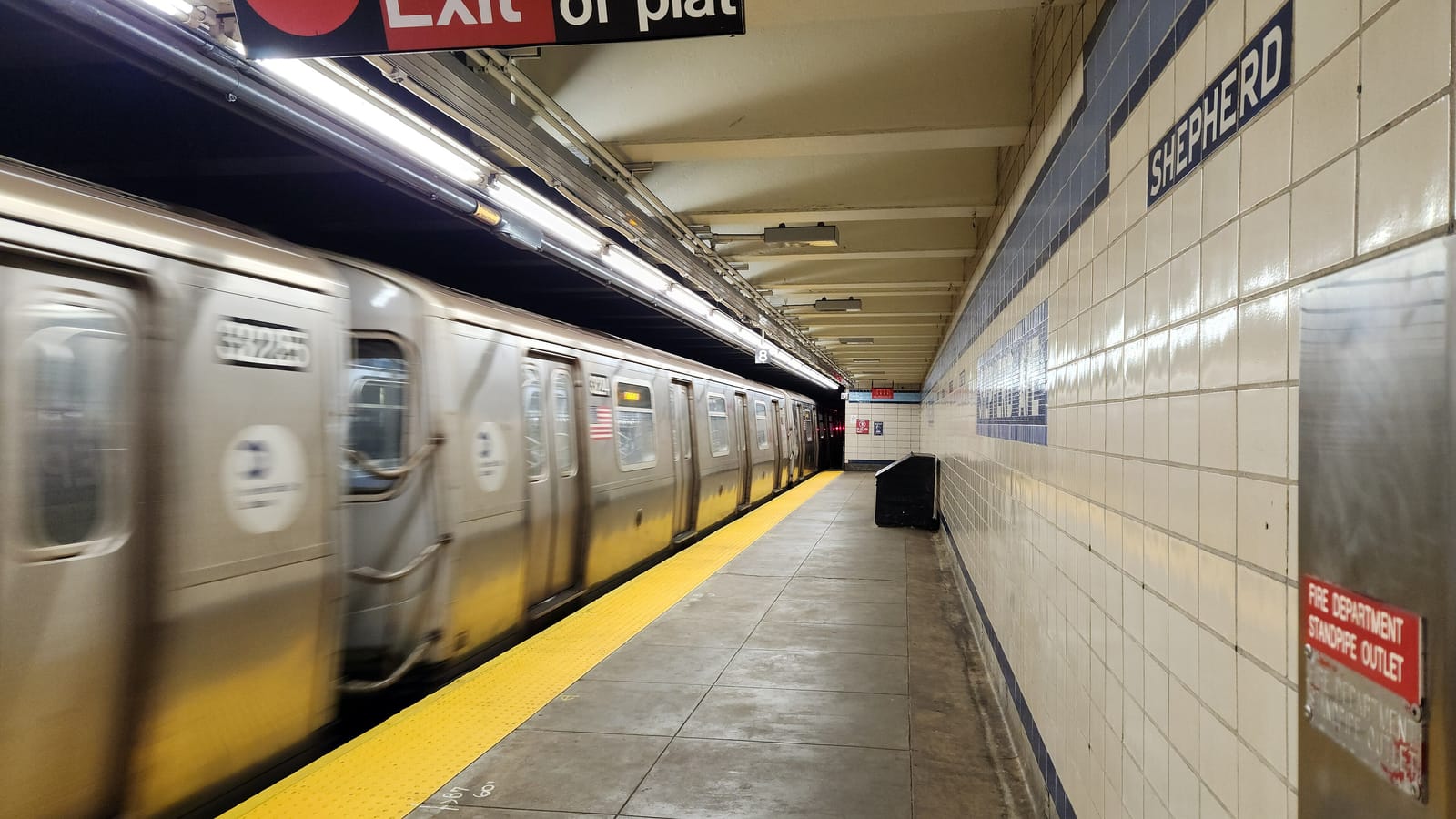 Photograph of a subway platform with tan and white wall tiles and a blue band reading Shepherd, a yellow platform edge, and a silver train blurred by motion along the left side, under a red exit sign