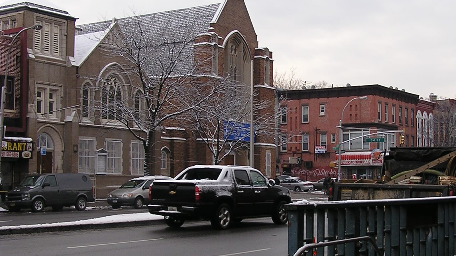 Photograph of a wide avenue in winter with light snow, a brick church with a pointed gable and tall arched windows, parked cars and a black pickup truck, red brick row buildings at a corner with a 54 St street sign, and the green railing of a subway entrance in the foreground