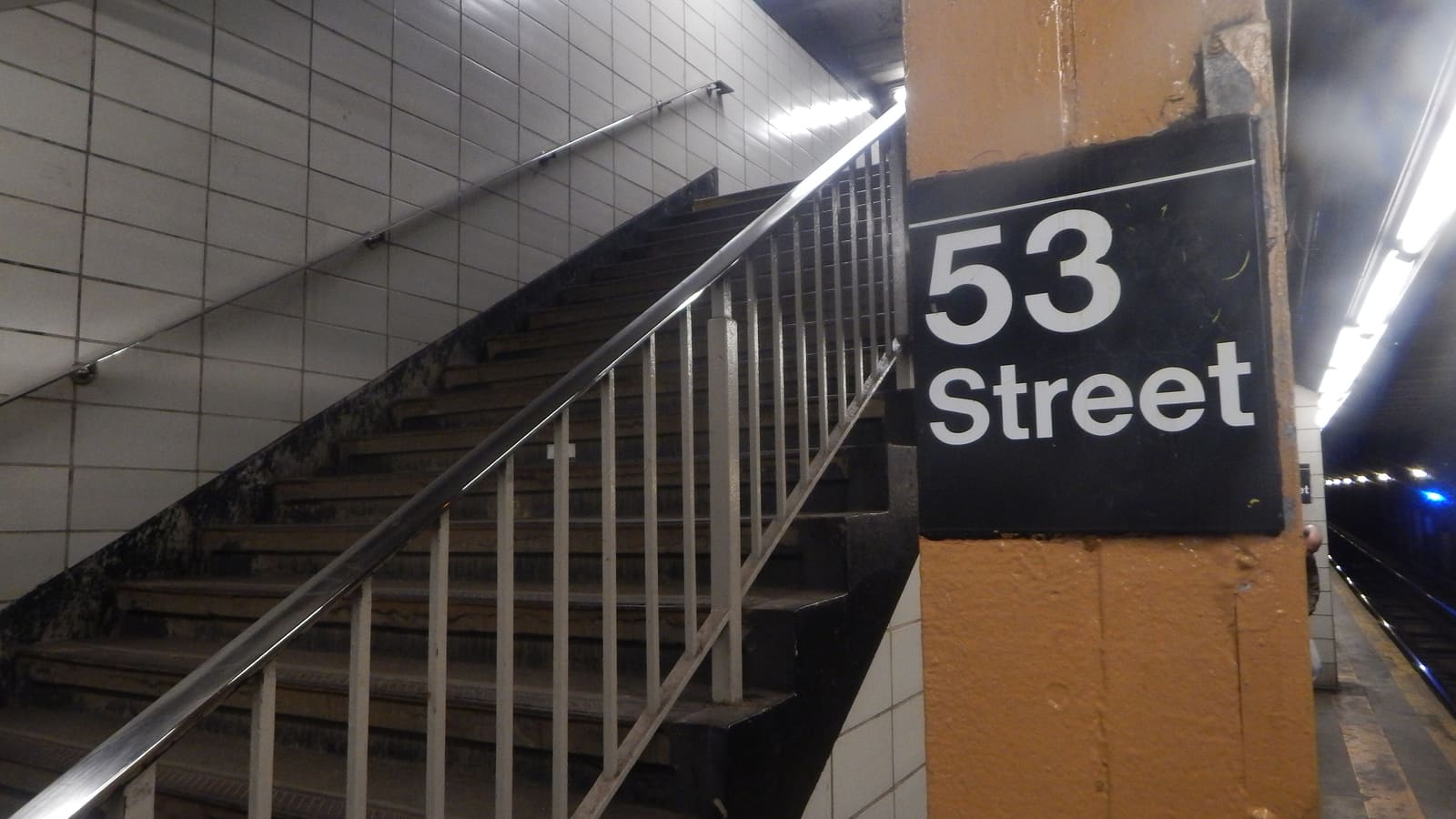 Photograph of a subway platform stair with a metal railing and white tile walls, beside an orange column with a black sign reading 53 Street