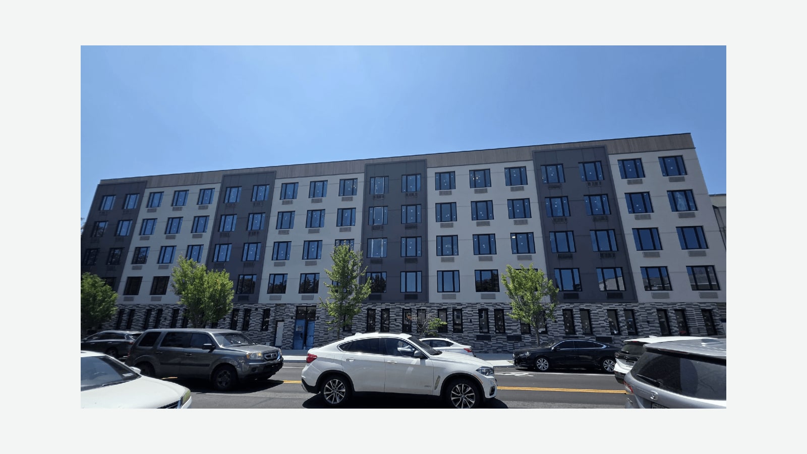 Photograph of a long six-story apartment building under a clear blue sky, with a dark patterned brick ground floor, five upper floors of pale gray and dark gray panels with rows of dark windows and wall air-conditioner grilles, a blue entrance door, young street trees, and parked cars along a street with a double yellow line, shown at its original size on a plain light background