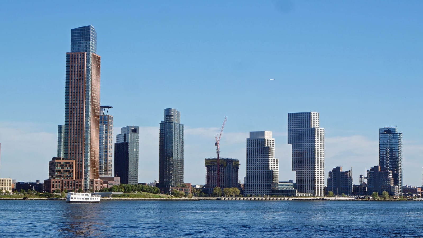 Photograph across the water of a waterfront skyline with several tall residential towers, one with a crane beside it, a ferry on the river and a low green park along the shore