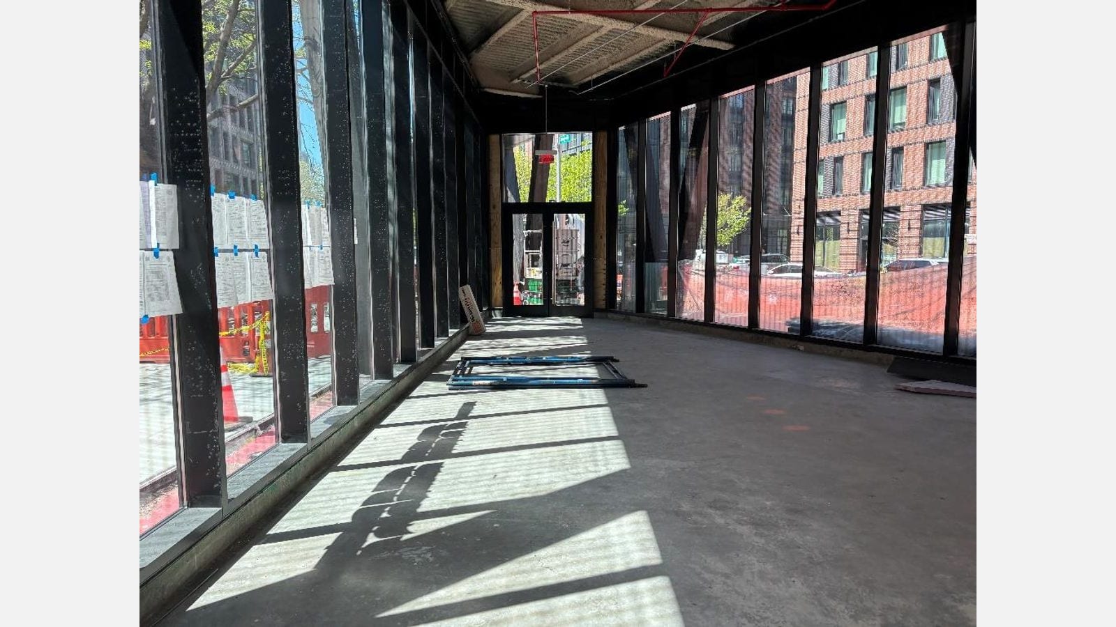 Photograph inside an unfinished ground-floor space with a bare concrete floor, tall black-framed glass walls on both sides with notices taped to the left wall, a glass door at the far end and red brick buildings across the street