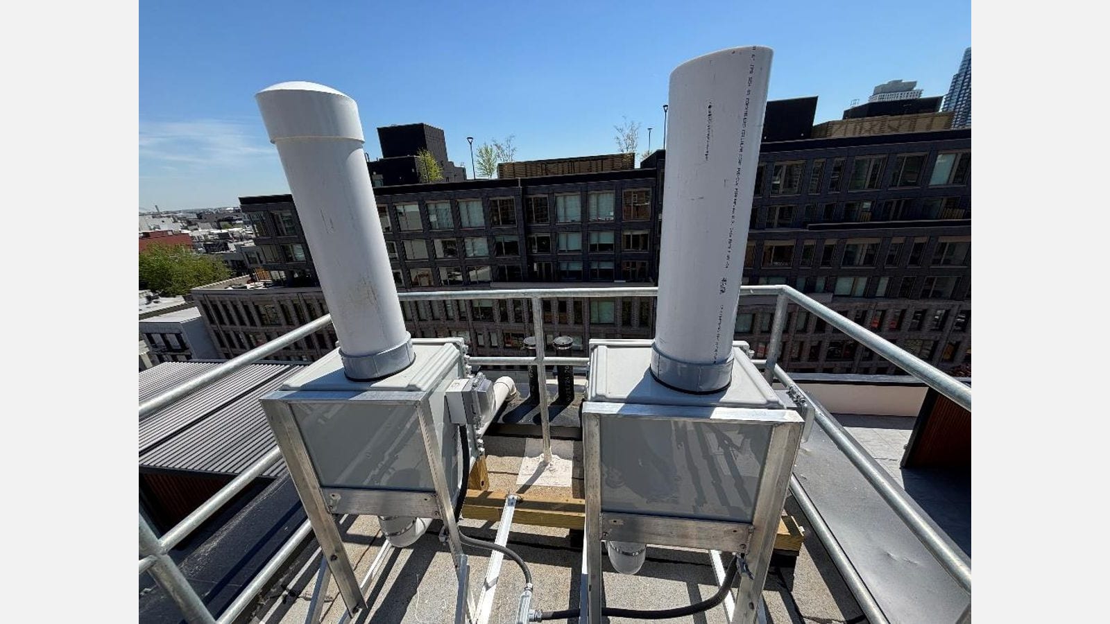Photograph of two white vent pipes rising from gray metal boxes on aluminum stands behind a pipe railing on a roof, with dark-brick apartment buildings and a clear blue sky behind