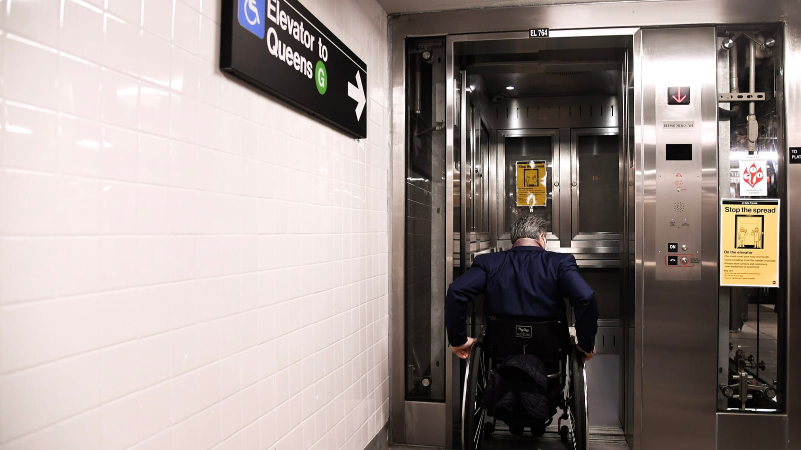Photograph in a white-tiled subway passage of a person in a wheelchair, seen from behind, rolling into an open steel elevator under a sign reading Elevator to Queens with a G train symbol