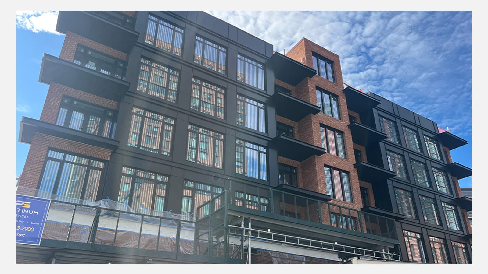 Photograph of a five-story building with red brick sections and black-framed window bays, black cantilevered balconies, a sidewalk shed and scaffold along the ground floor, and a contractor's blue banner at the left, under a blue sky with clouds