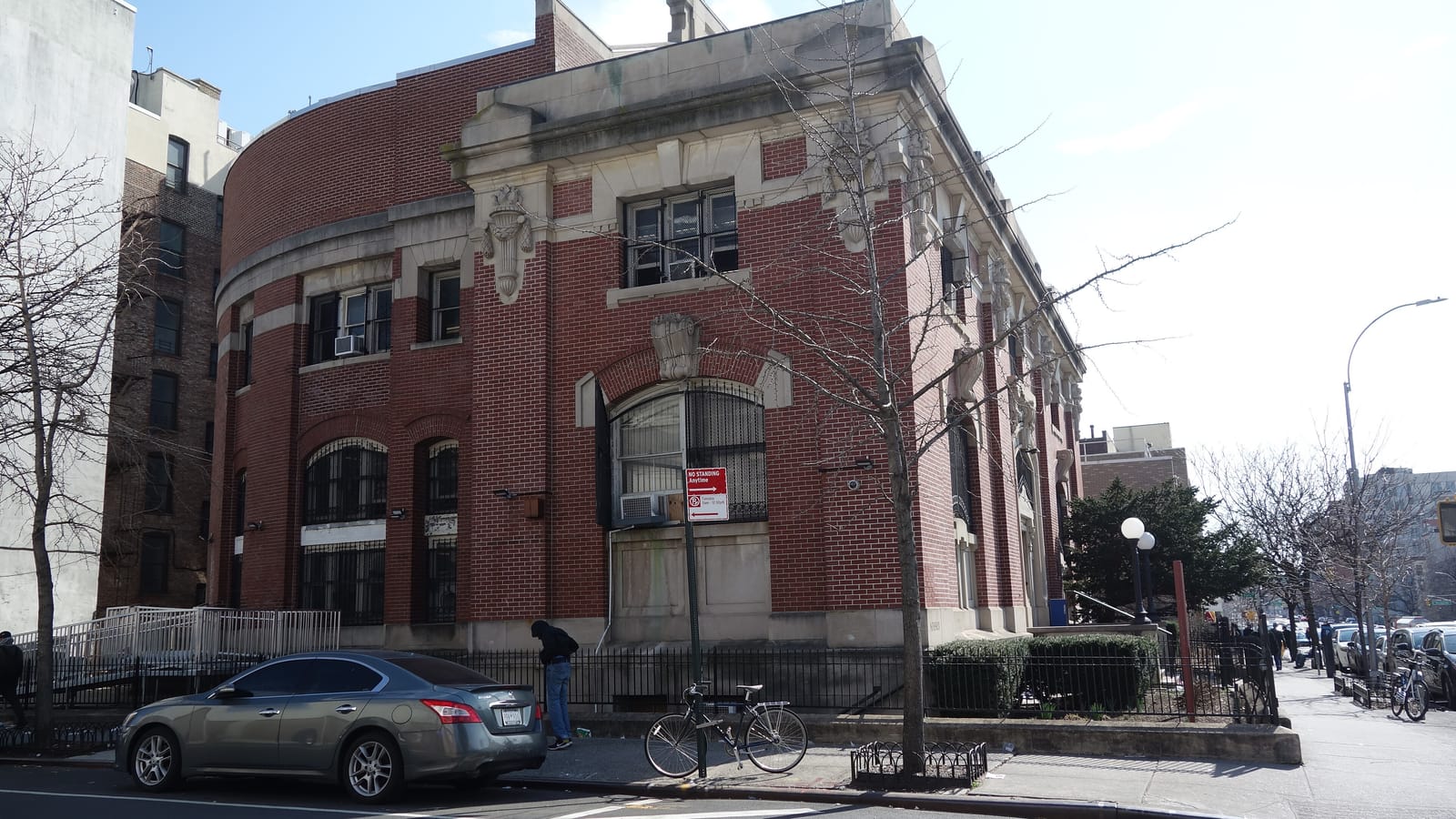 A two-story red brick library with pale stone trim, carved urns and tall barred arched windows, a rounded rear wing, a bare street tree and a no-standing sign on the sidewalk, a gray parked car and a bicycle locked to a pole in front