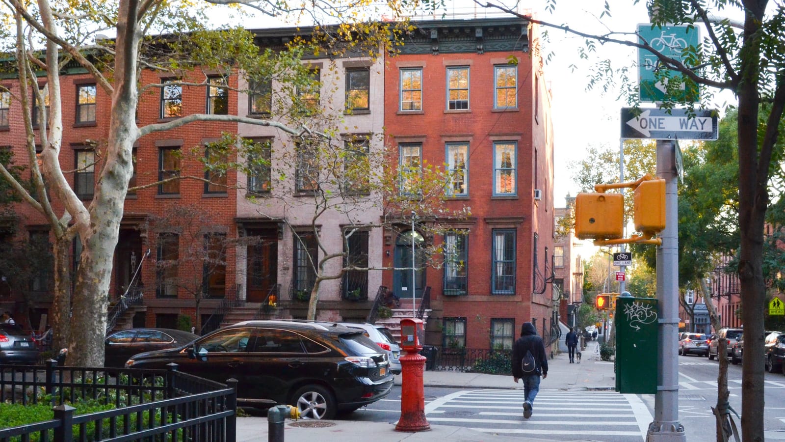 A row of four-story red and pink brick rowhouses with stoops behind street trees in autumn, a red fire alarm post and a black car at the curb, a one-way sign and pedestrian signal on a pole, and a person walking away across a crosswalk