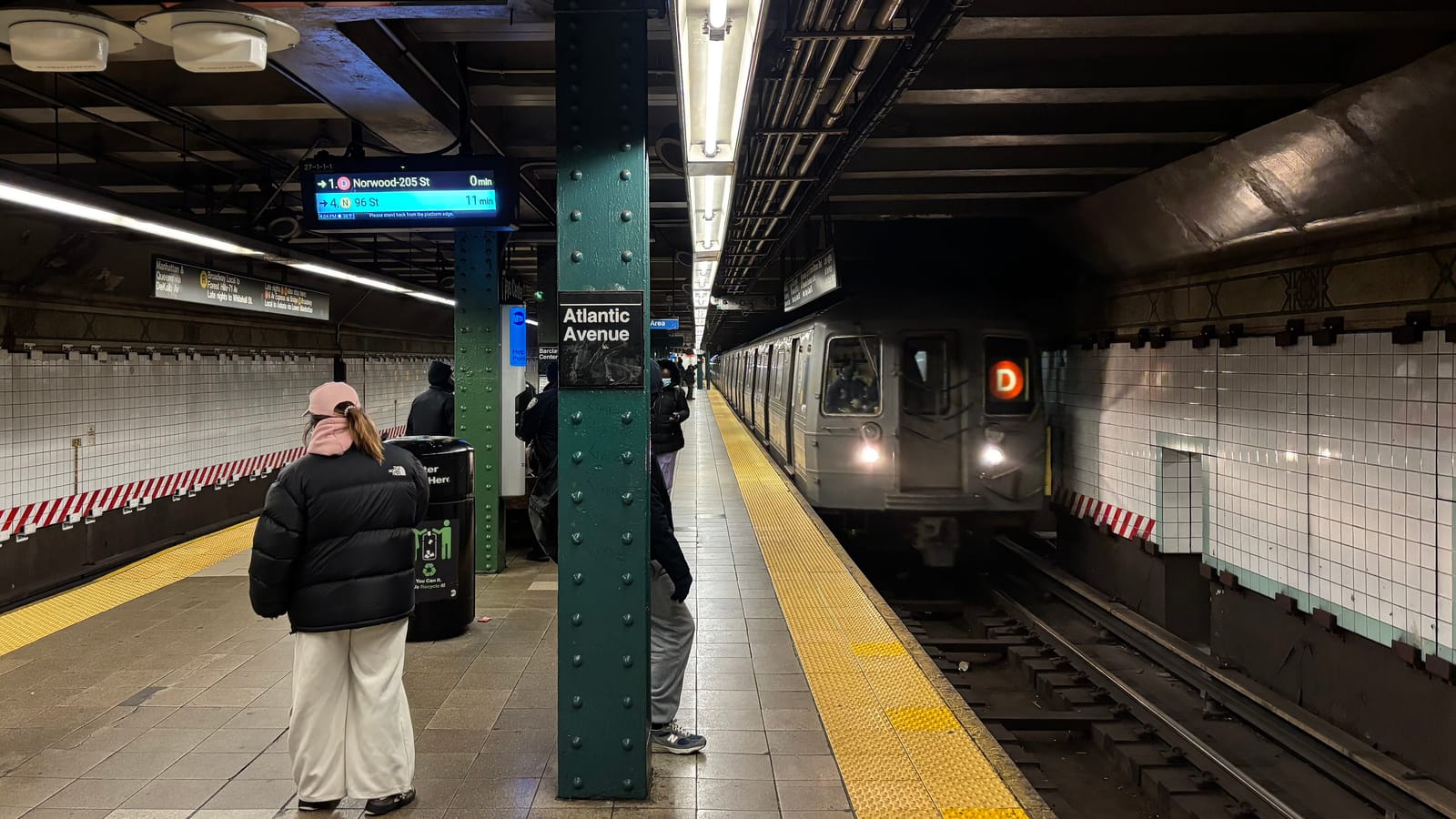 A subway platform with a green riveted column marked Atlantic Avenue, a yellow tactile strip along the edge, a D train arriving with its headlights on, a countdown sign above, and a few riders waiting, seen from behind