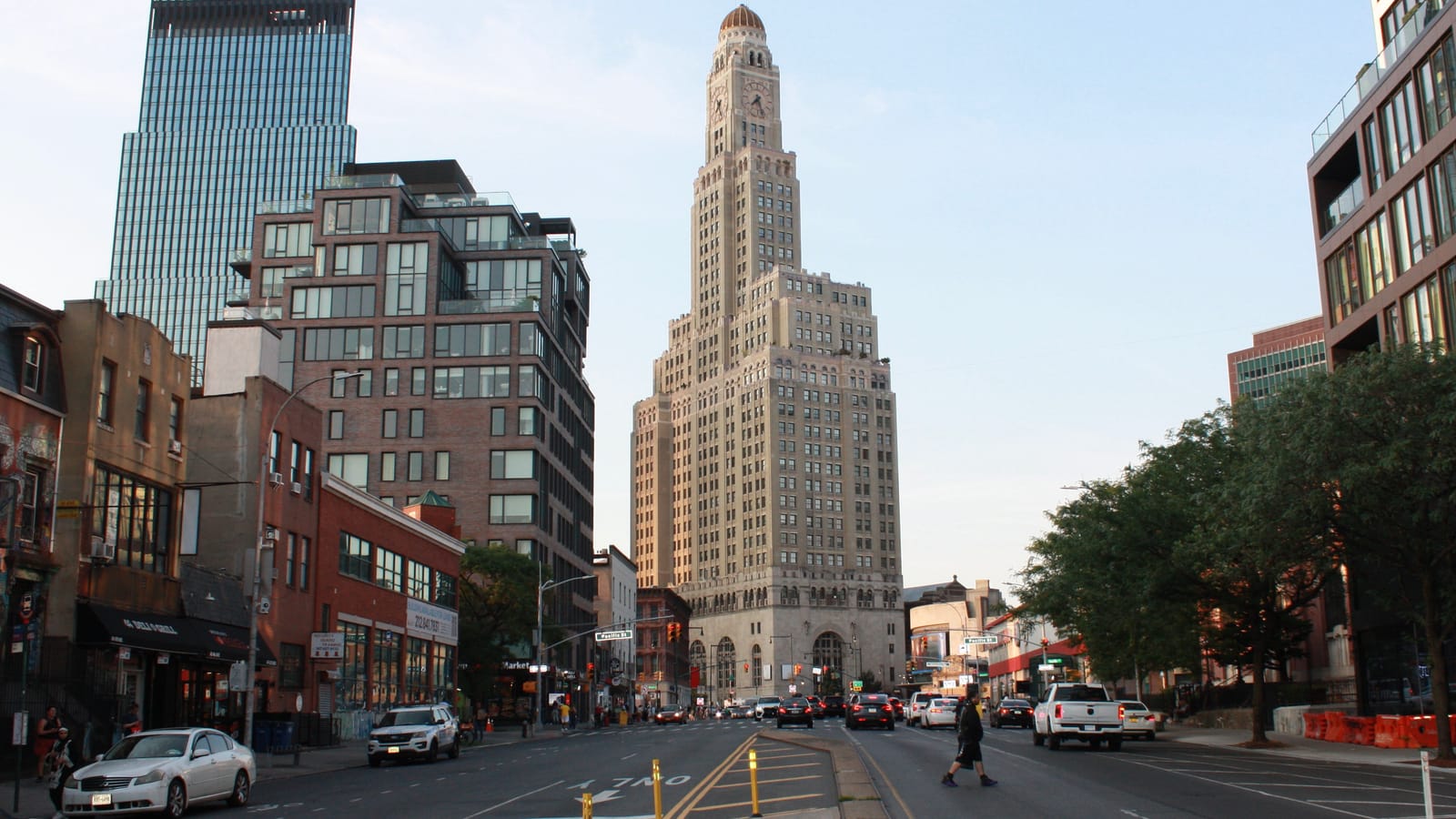 A wide avenue at dusk looking north toward a tall stone clock tower with a gold dome, with a dark glass skyscraper and a brick apartment building on the left, low shops and parked cars along the street, trees on the right, and one person crossing the road far ahead