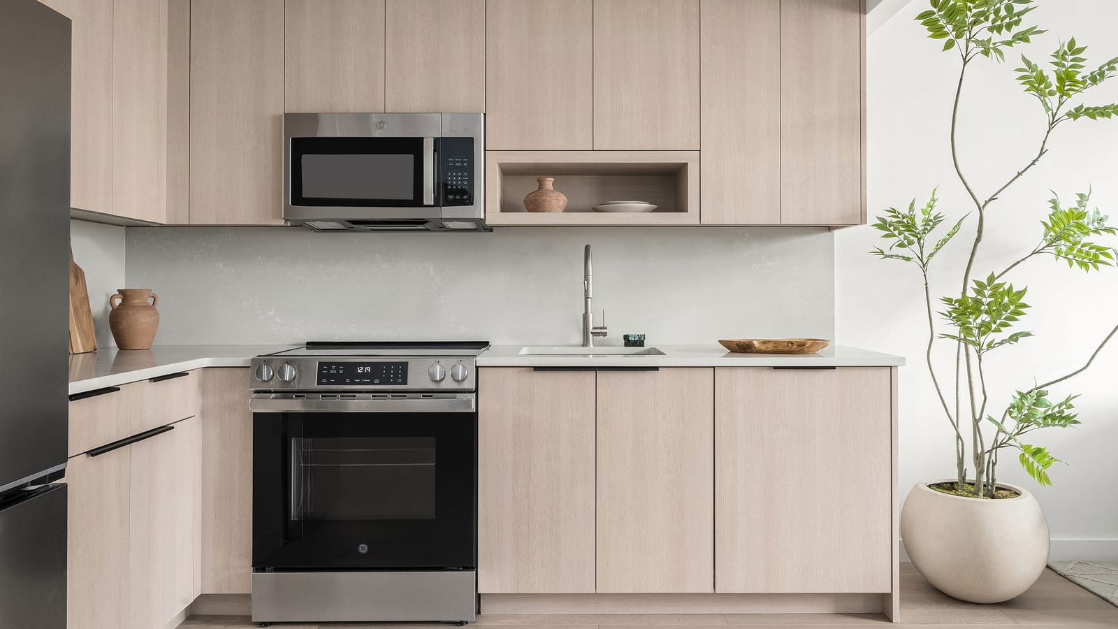 Photograph of a kitchen wall with pale wood-grain cabinets, a white stone counter and backsplash, a stainless-steel microwave over a stainless-steel GE range with a smooth black glass cooktop, a sink with a tall faucet, a refrigerator edge at the left and a tall potted plant at the right