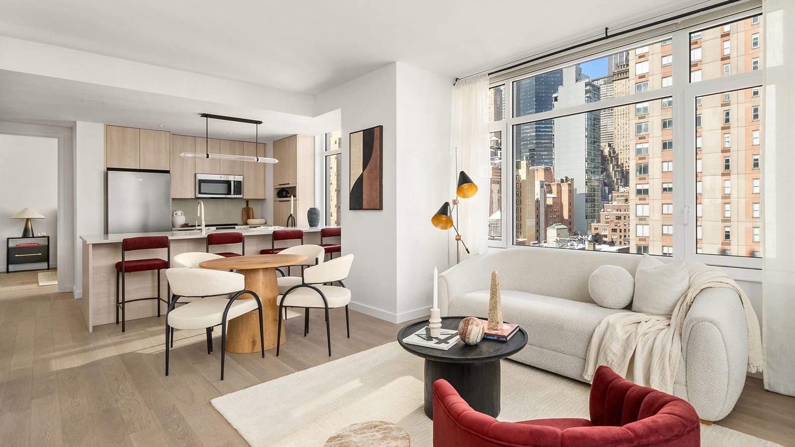 Photograph of a furnished living and dining room with light wood floors, a white sofa, a red armchair and a round dark coffee table near a wide corner window looking at Midtown towers, a round wood dining table with four chairs, and an open kitchen behind a counter with four red stools