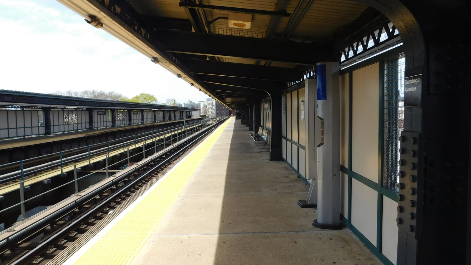 Photograph of an empty elevated subway platform under a dark steel canopy, with a yellow edge strip, tracks at left, a steel column with a sign reading Saratoga Avenue at right, and apartment buildings in the distance