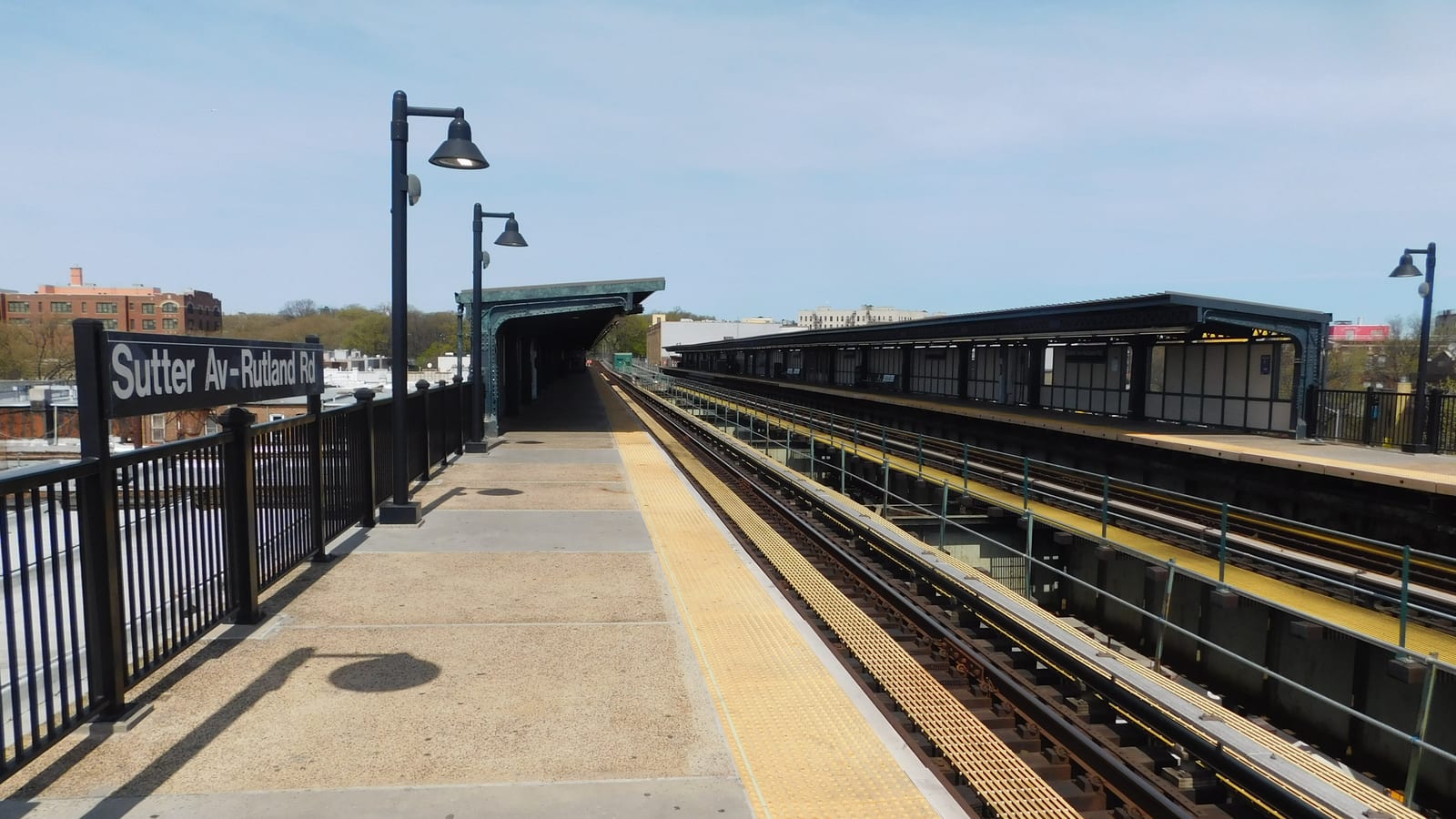 Photograph of an open elevated subway platform with a black sign reading Sutter Av-Rutland Rd on a railing, black lamp posts, a yellow edge strip, tracks and a second covered platform at right, and low brick buildings beyond