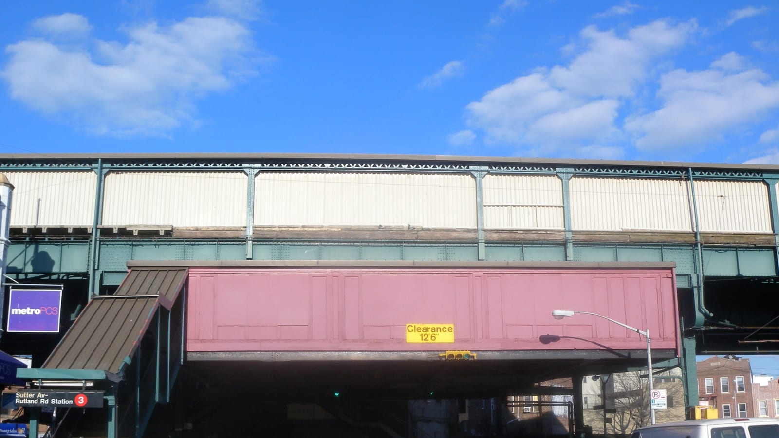 Photograph of an elevated subway structure with a cream-colored wall and a pink enclosed section marked Clearance 12 feet 6 inches, a covered street stair at left under a sign reading Sutter Av-Rutland Rd Station and a red 3, under a blue sky with clouds