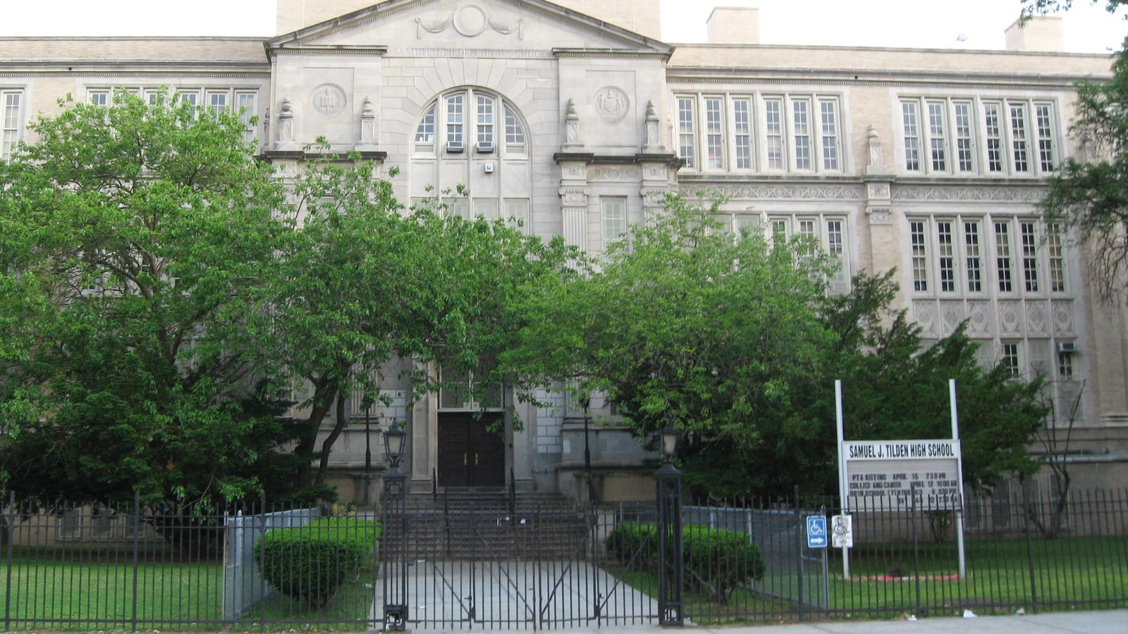 Photograph of a large pale stone school building with an arched central window and a carved pediment, trees in front, a black iron fence and gate, and a sign reading Samuel J. Tilden High School on the lawn at right