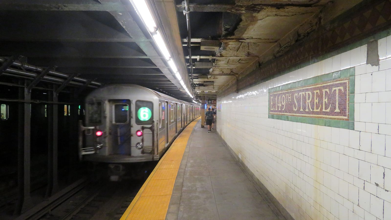 An underground subway platform with white tiled walls and a mosaic name sign reading E. 149th Street, a 6 train pulling away on the left and two people walking far down the platform