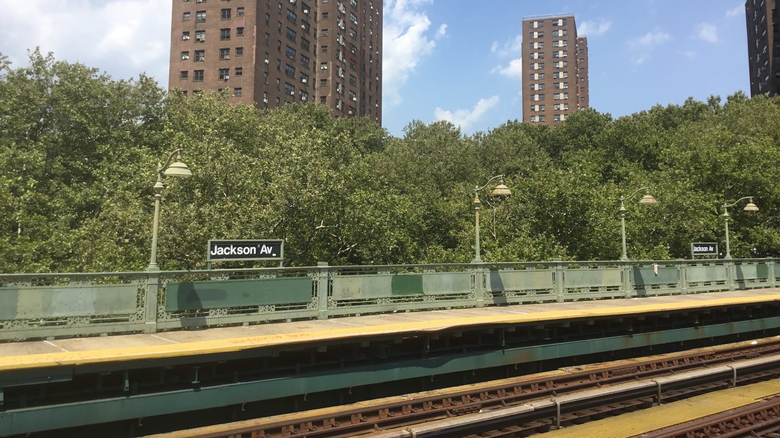 An outdoor subway platform with a green iron railing, old-style lamp posts and a sign reading Jackson Av, with tracks in front, tall trees behind and tall brick apartment towers above the trees