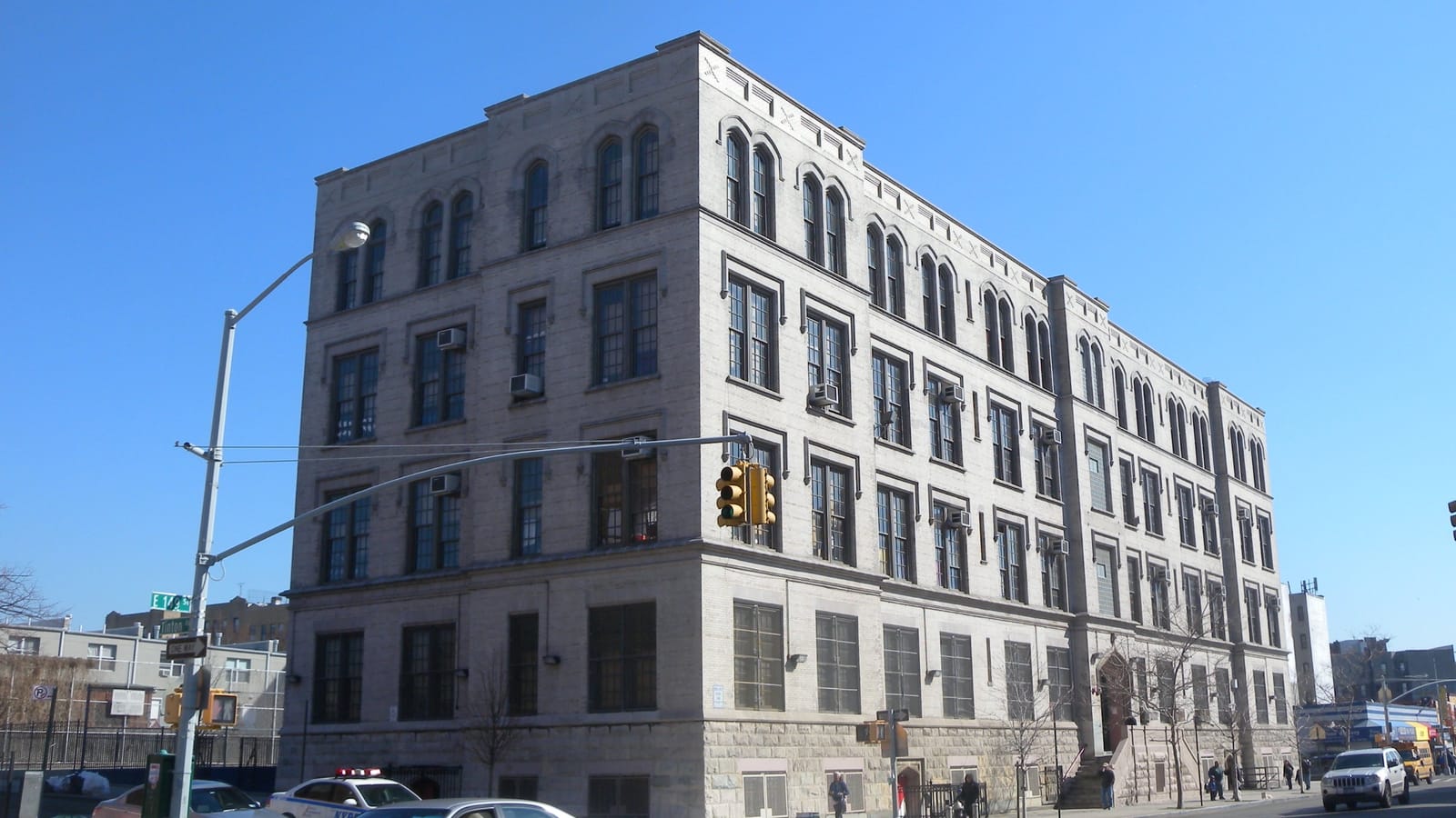 A four-story gray stone school building with arched top-floor windows on a street corner, a traffic signal and a street sign reading E 149 St at left, a police car and parked cars in front, under a clear blue sky