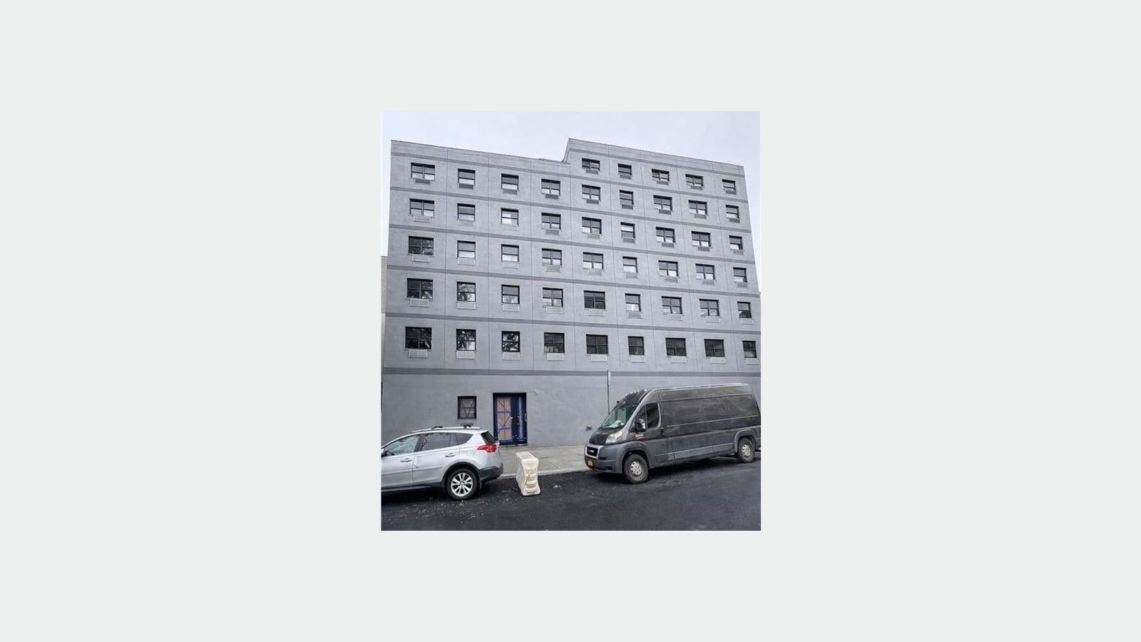 Photograph of a plain gray seven-story apartment building with rows of square black windows and wall vents under each window, a front door covered with protective board, and a silver SUV and a dark van parked at the curb in front