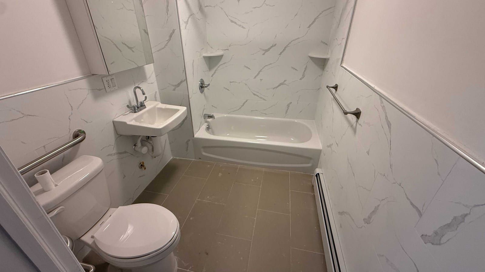 A bathroom with white marble-look tiles on the walls: a white bathtub with a shower head and corner shelves, a wall-hung sink with a lever faucet and a mirrored cabinet above it, a toilet with a steel grab bar on the wall beside it, a towel bar, a baseboard heater and brown floor tiles