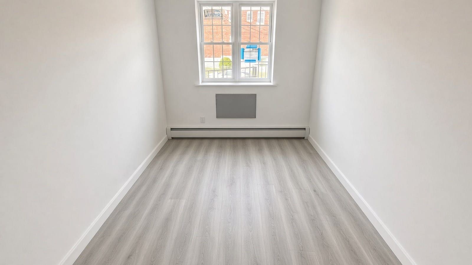 A narrow, empty room with white walls and light grey wood-look flooring, a white baseboard heater along the far wall under a double window, and a grey wall-sleeve cover below the window; through the window, a red brick building and a railing outside