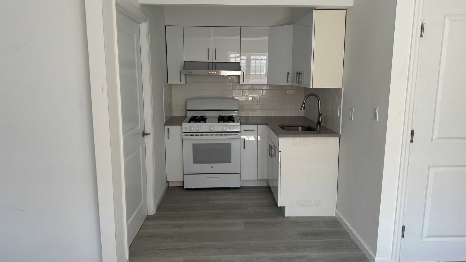 A small kitchen set into an alcove: glossy white upper and lower cabinets, a white subway-tile backsplash, a white GE gas range with four burners under a stainless range hood, a grey counter and a stainless sink with a tall faucet, with white doors on both sides and grey wood-look flooring