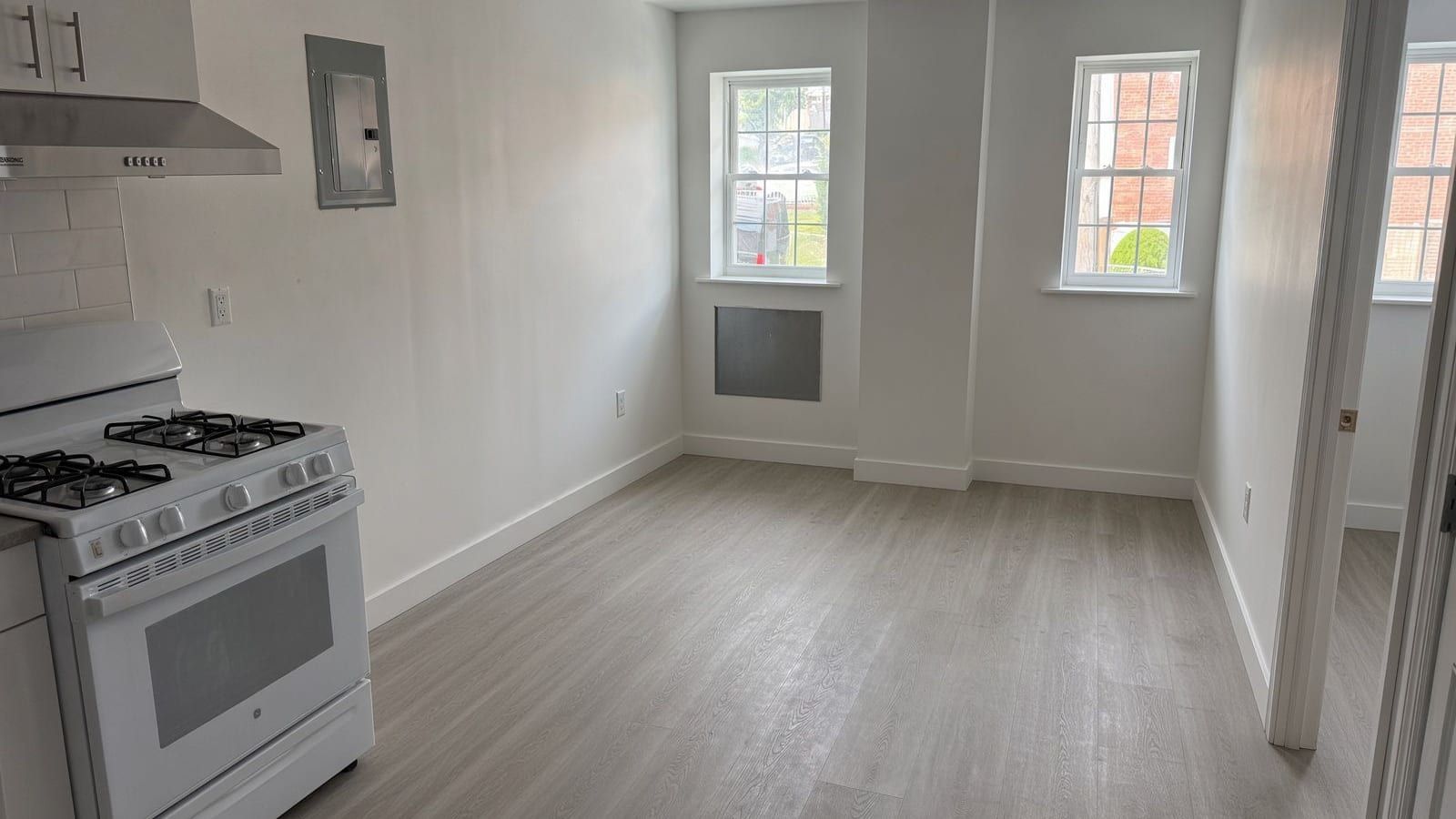 An empty room with pale grey wood-look flooring and white walls: a white four-burner gas range with a stainless range hood and an electrical panel on the wall at left, two double-hung windows with grey wall-sleeve covers below one of them at the back, and an open door to another room with a window at right