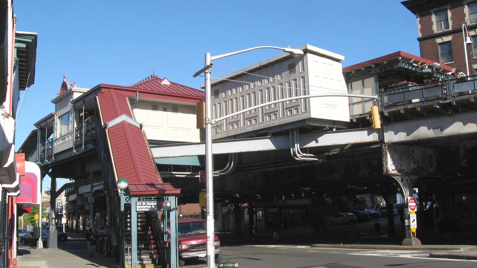 Photograph of an elevated subway station over a street corner, with a red-roofed station house, a covered red stair down to a green entrance kiosk with a sign for the 174 St station and the 2 and 5 trains, a street lamp, a parked red van and the steel structure of the elevated line