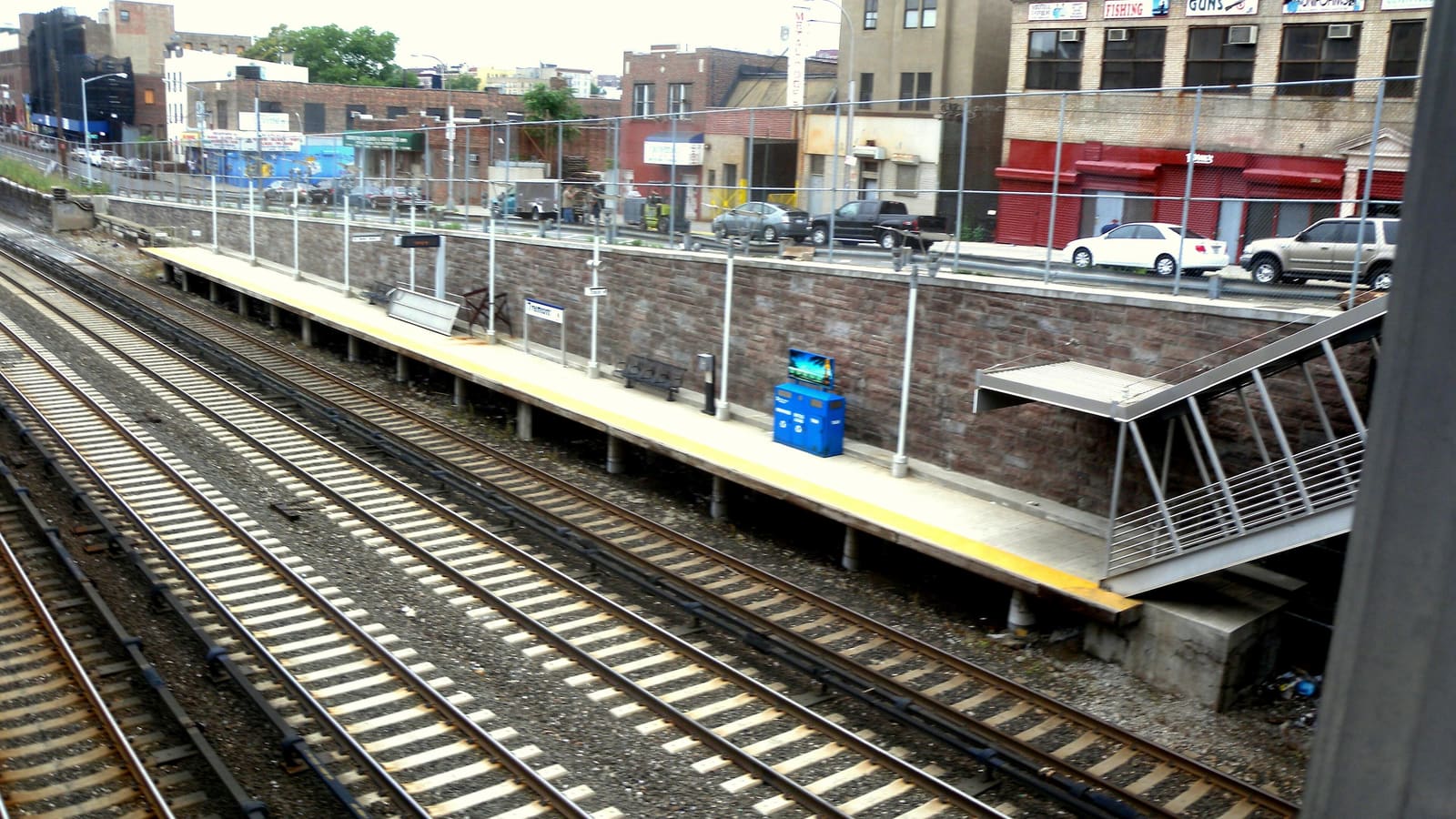 Photograph of a Metro-North platform seen from above, with four tracks in the foreground, a long platform with a yellow edge, a blue bin and benches against a stone wall, a metal stair at right, and older commercial buildings and parked cars on the street above