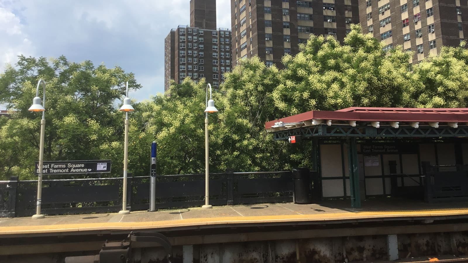 Photograph of an elevated subway platform with a sign reading West Farms Square East Tremont Avenue, white lamp posts, a dark railing, a canopy with a red roof at right, leafy trees behind and tall brown brick apartment towers against a cloudy sky