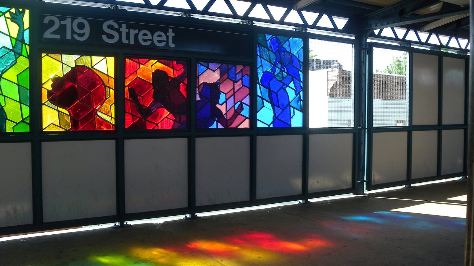 Photograph of an elevated subway platform windbreak with bright stained-glass panels in red, yellow, green, purple and blue showing figures, under a black sign reading 219 Street, with sunlight throwing colored light across the platform floor