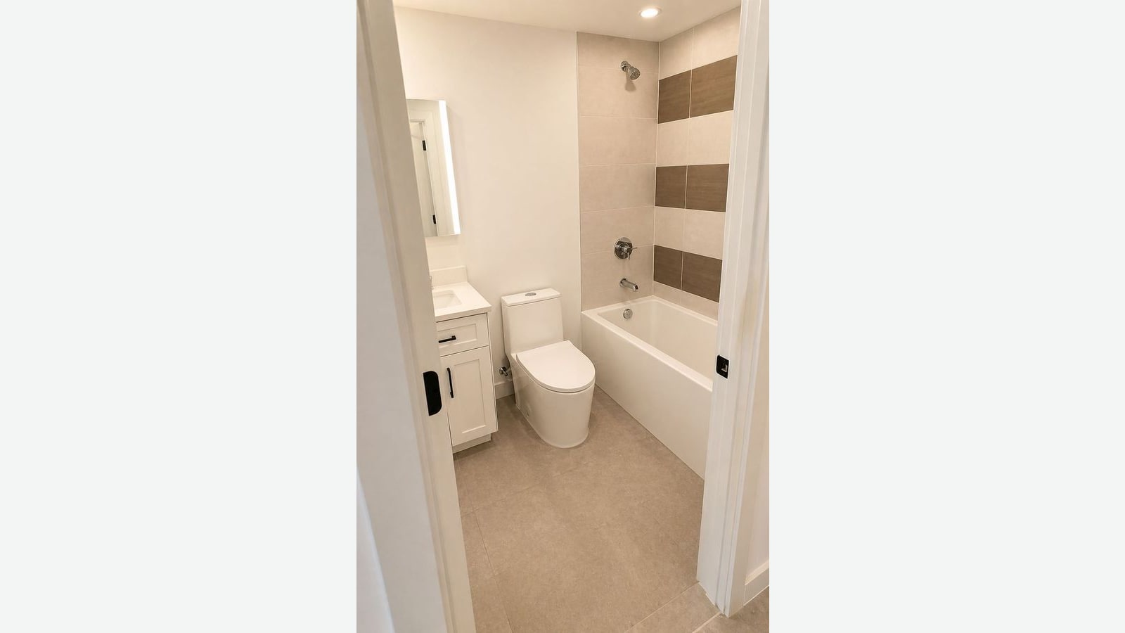 Photograph through an open door into a bathroom with beige floor tile, a white vanity with a black handle and a lit mirror cabinet, a white one-piece toilet, and a white tub with a chrome shower set against beige and brown striped wall tile