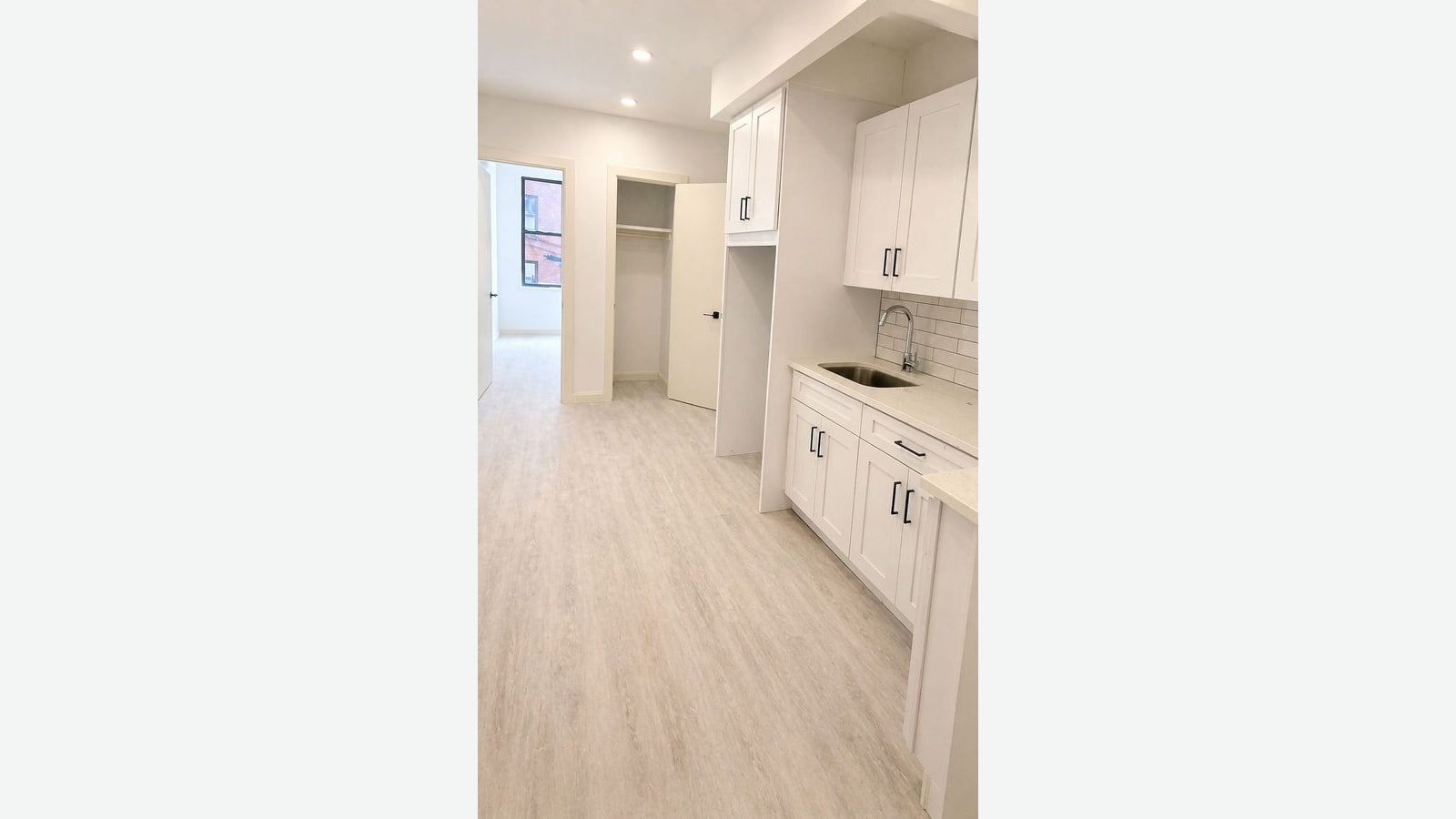 Photograph of a narrow white room with light wood-look vinyl flooring, white shaker kitchen cabinets with black handles along the right wall, a stainless sink with a gooseneck faucet under white subway tile, an open closet with a shelf and rod, and a doorway at the back into a bright room with a window facing a brick building