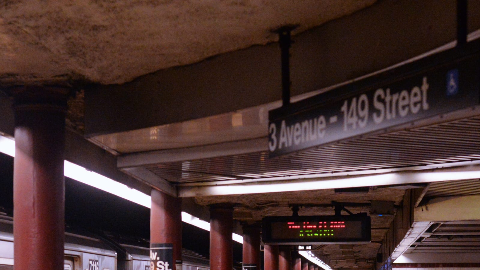Photograph of an underground subway platform with red columns, a hanging sign reading 3 Avenue - 149 Street with a wheelchair symbol, a red and green arrival display, column signs reading 149 St and the side of a silver train at lower left