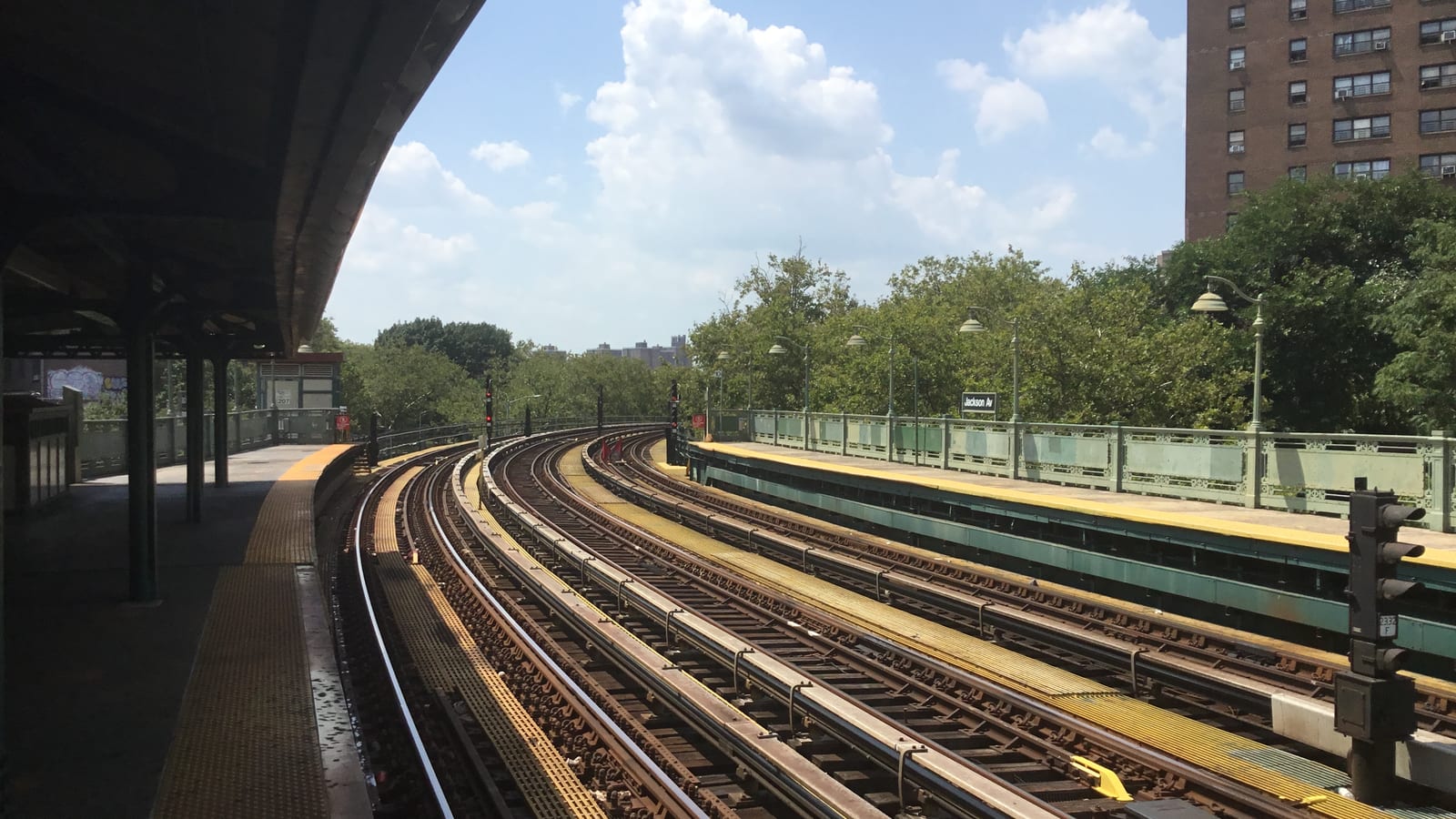 Photograph of four curving subway tracks between two outdoor platforms, with a platform canopy at left, a Jackson Av sign and lamp posts along a green railing at right, trees and a tall brown brick tower behind, under a blue sky with large white clouds