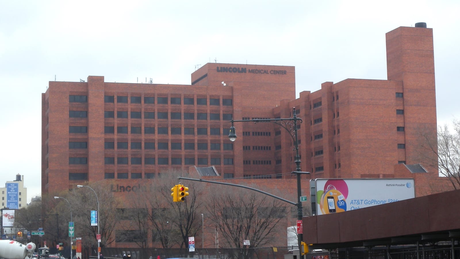 Photograph of a large red-brick hospital building with lettering reading Lincoln Medical Center near the roof, a tall brick tower at right, bare trees, a traffic light and street lamp in front and a billboard at lower right, under a gray sky