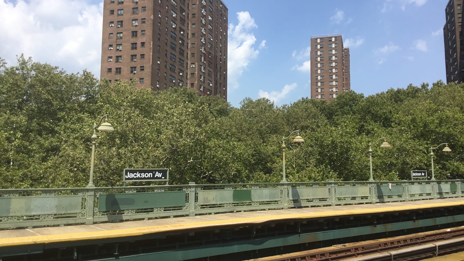 Photograph from a subway platform above the street, with a black station sign reading Jackson Av on a green iron railing, old-style lamp posts, a dense row of leafy trees behind and three tall brown brick apartment towers against a blue sky with white clouds