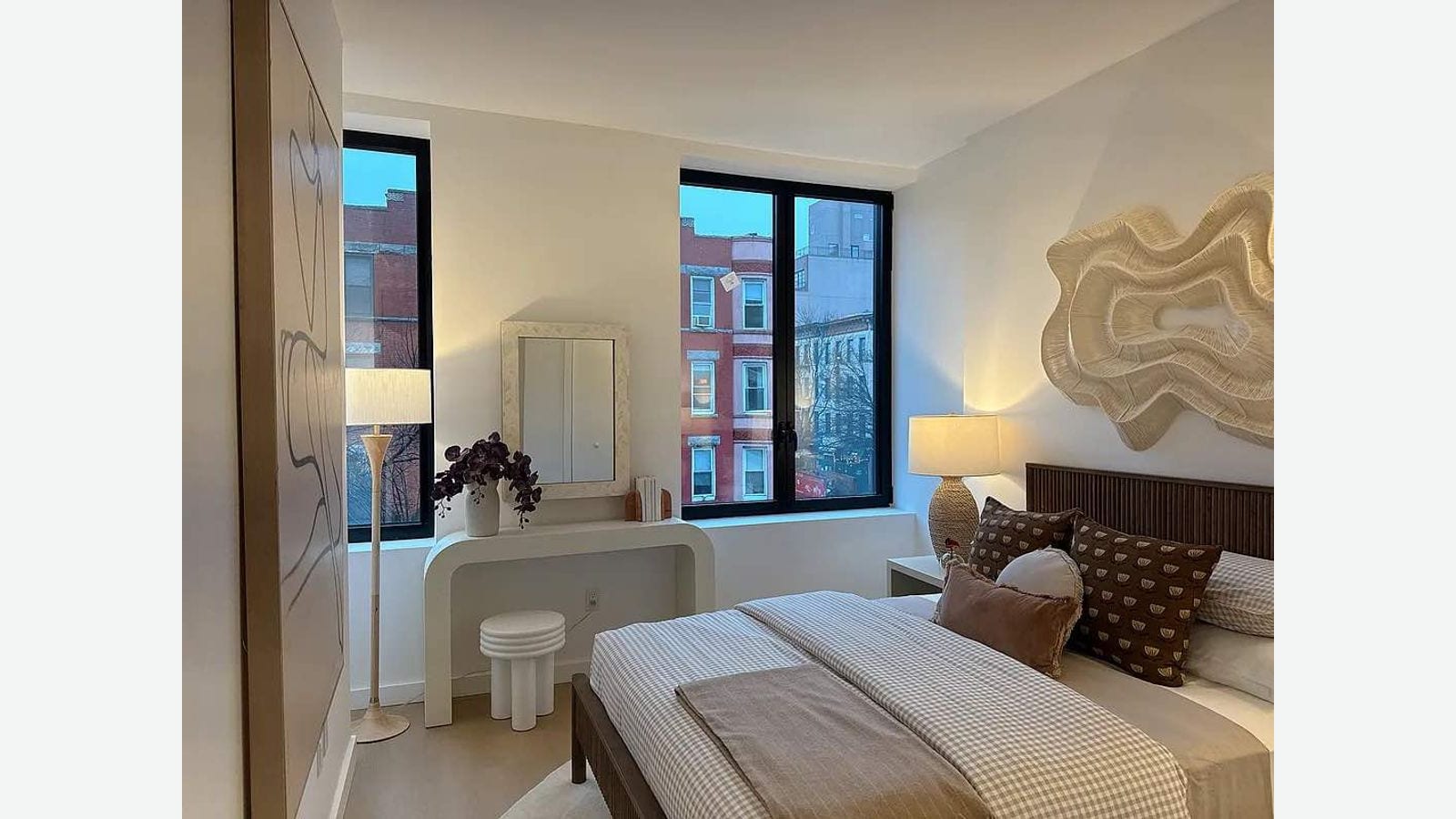 Photograph of a bedroom with two black-framed windows facing brick row houses at dusk, a bed with checked bedding and patterned pillows, a lamp on a nightstand, a white vanity with a mirror and a sculpted wall hanging